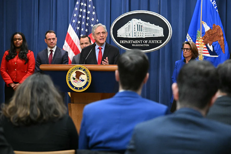 A gray haired man speaks at a podium in front of a US flag and several other people.