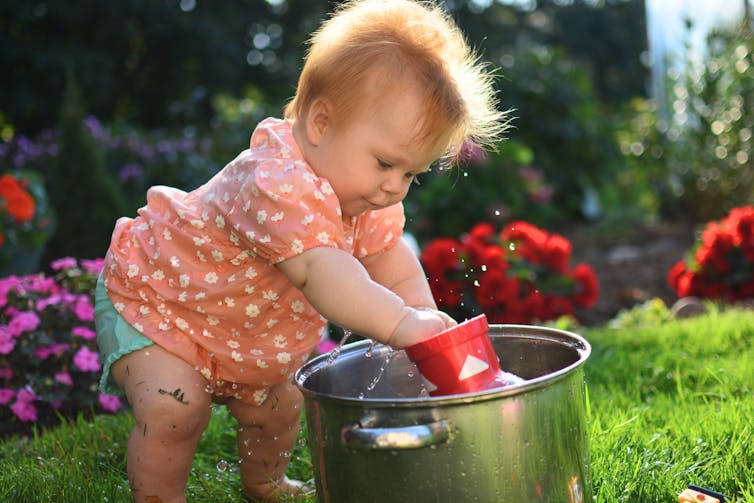 Baby girl in pink dress is playing with water in pot.