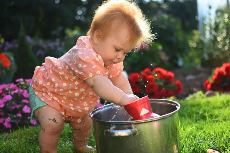 Baby girl in pink dress is playing with water in pot.