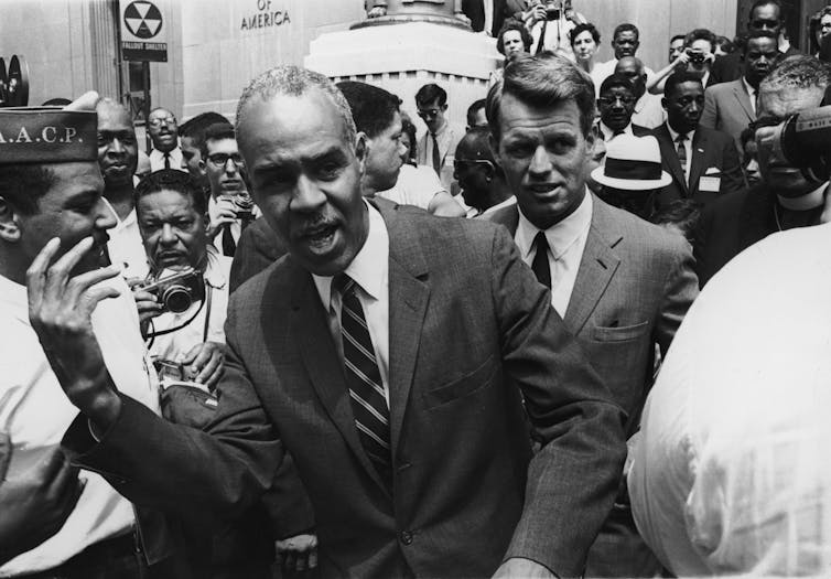 Two men in suits walking through a crowd outside.