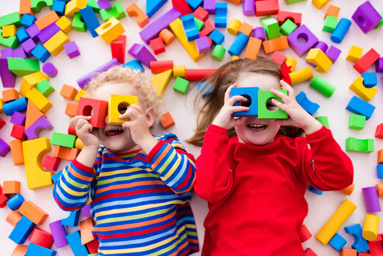 Two young children lying on floor covered with coloured blocks, holding some blocks over their eyes.