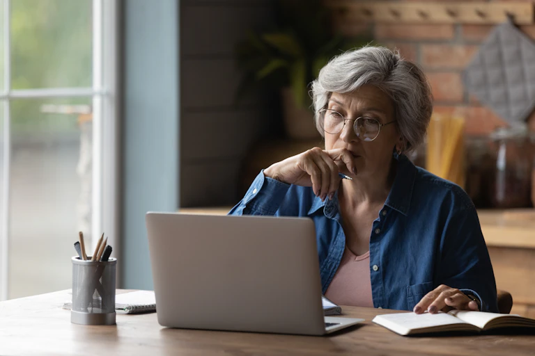 an older woman with grey hair, sitting at a desk working on a laptop