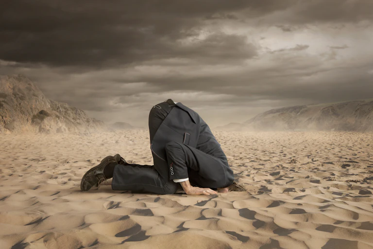 A person wearing a dark suit sticks his head in the sand.