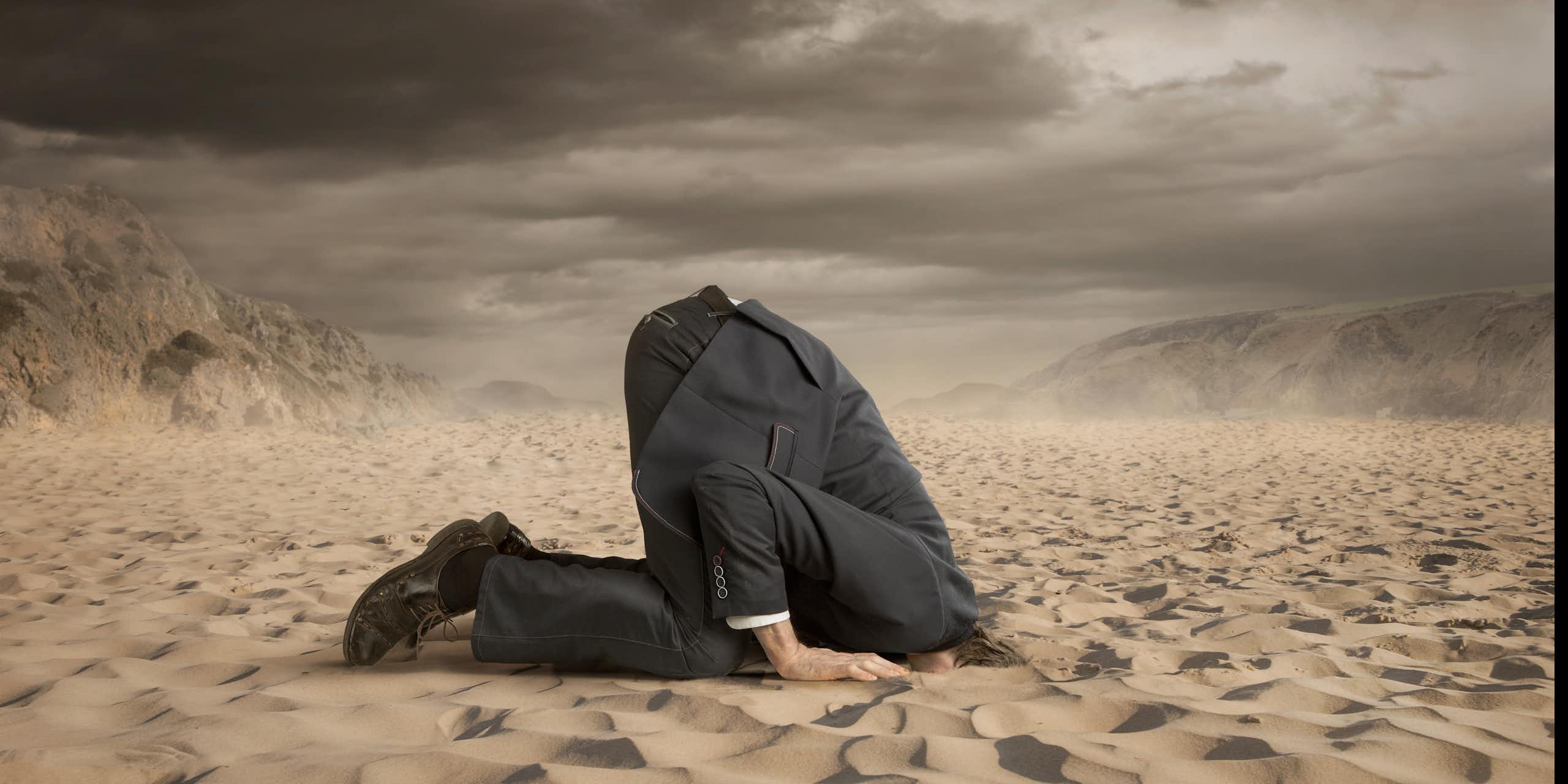 A person wearing a dark suit sticks his head in the sand.