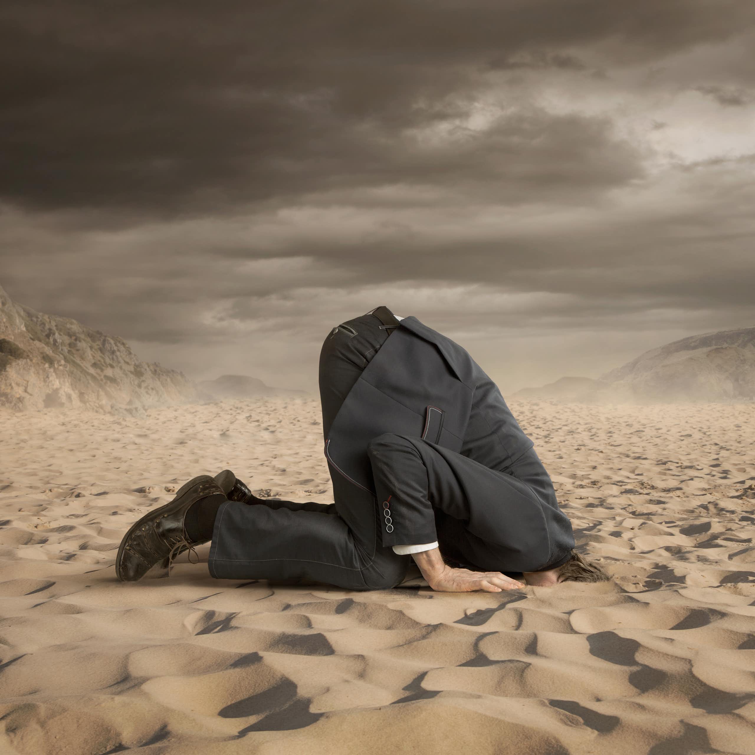 A person wearing a dark suit sticks his head in the sand.