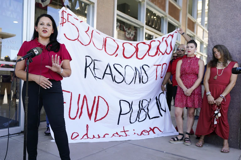 a woman wearing red speaks in front of a microphone in front of a sign that reads $5,300,000,000 reasons to fund public education