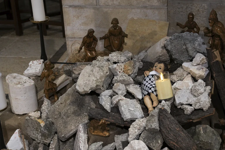 Figure of a baby wrapped in a keffiyah amid rubble with a candle in front and statue figures behind.