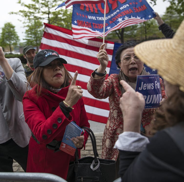 Two women wearing red stand in front of a crowd of people and an American flag and point their finger at someone.