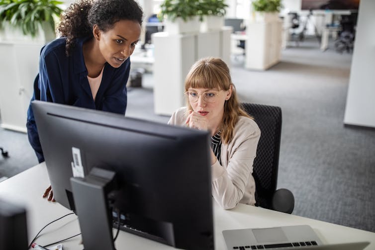 two women, one seated and one standing, look at a computer monitor