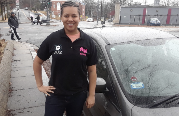 A smiling professor stands in front of a car, her hand on her hip. She is dressed all in black, and her shirt has the Uber and Lyft logos.