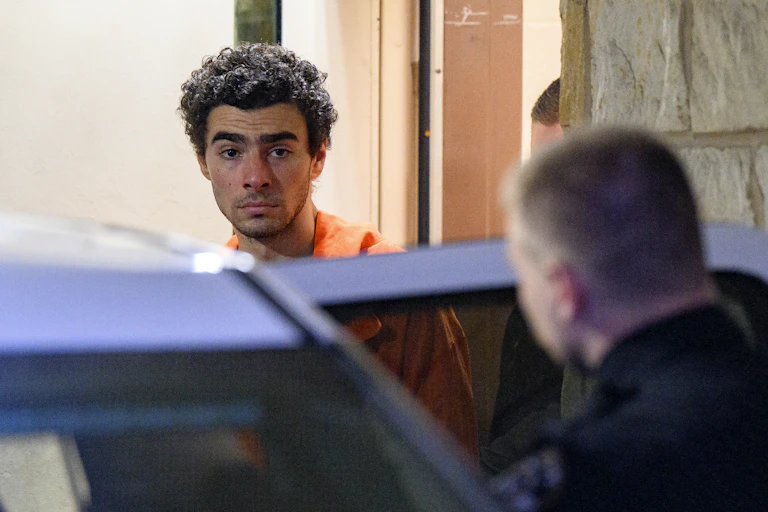 Young man with curly hair wears an orange jumpsuit as he is escorted into a car.