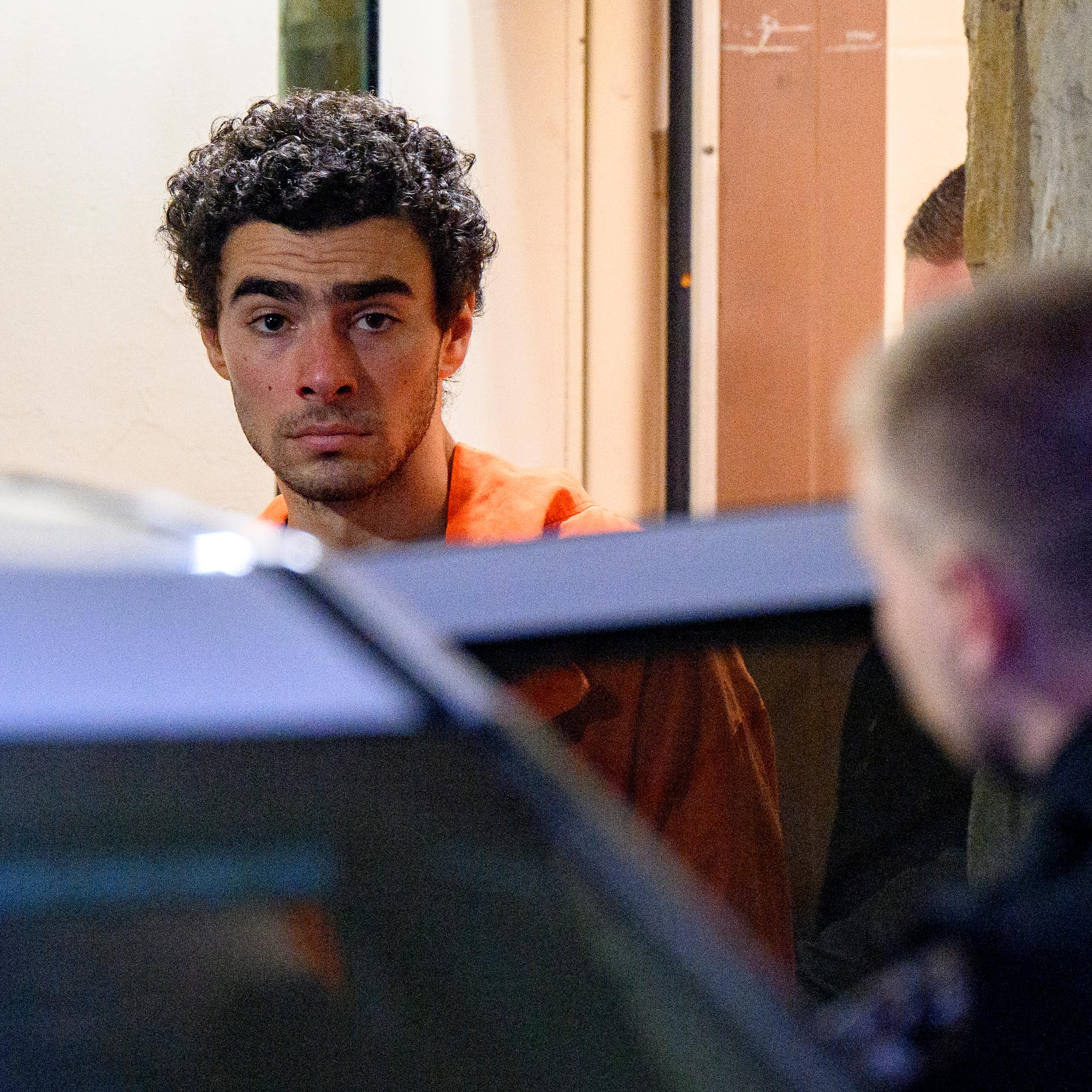 Young man with curly hair wears an orange jumpsuit as he is escorted into a car.