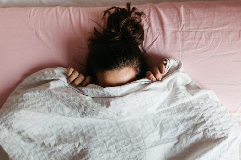 Young woman in bed, white bedcover up to her eyes, head sticking out against pink pillow