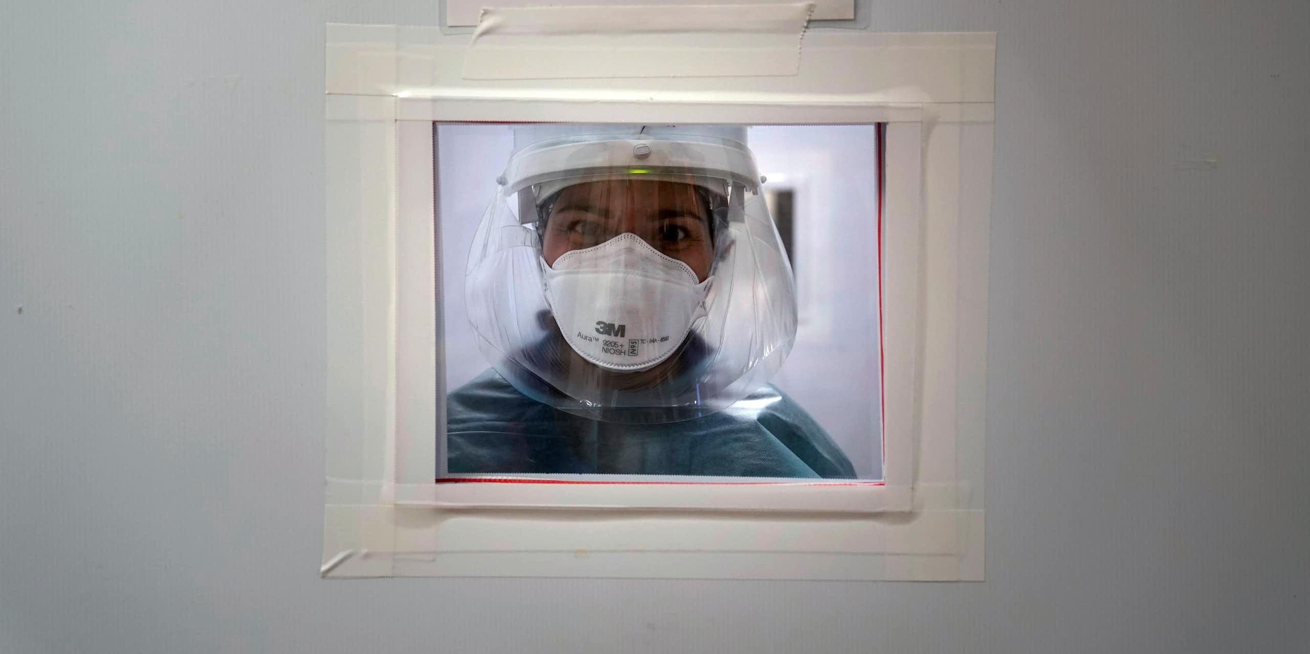 A woman in medical protective equipment peers through a small window cut into a white door.
