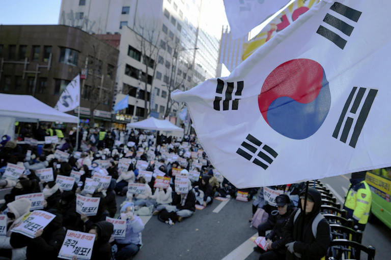 Photo of a a group of protesters sitting in a city street with a South Korean flag in the foreground.