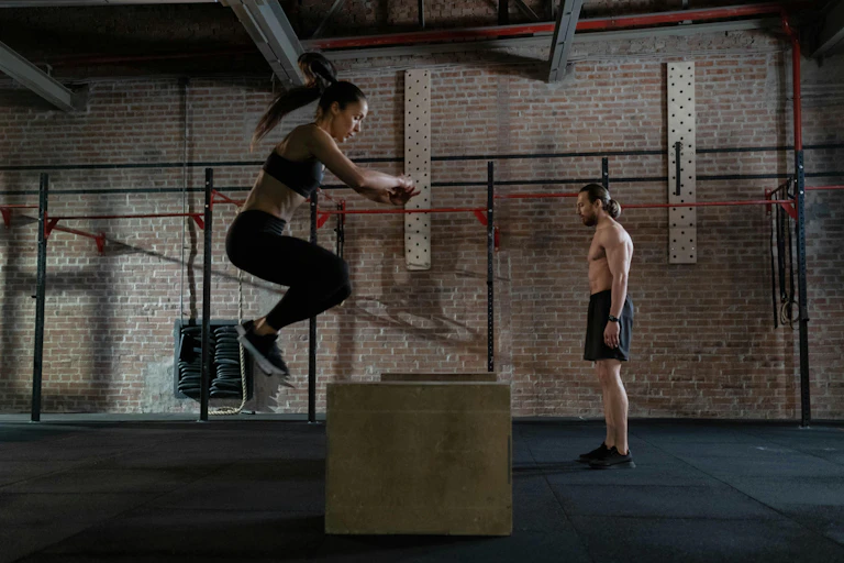 A man and a woman do box jumps in a gym.