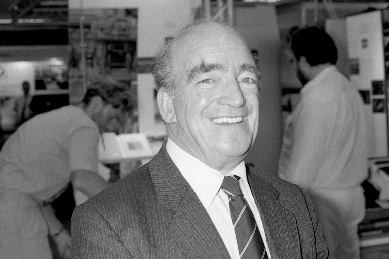 Smiling man in suit and tie, with factory workers behind him.