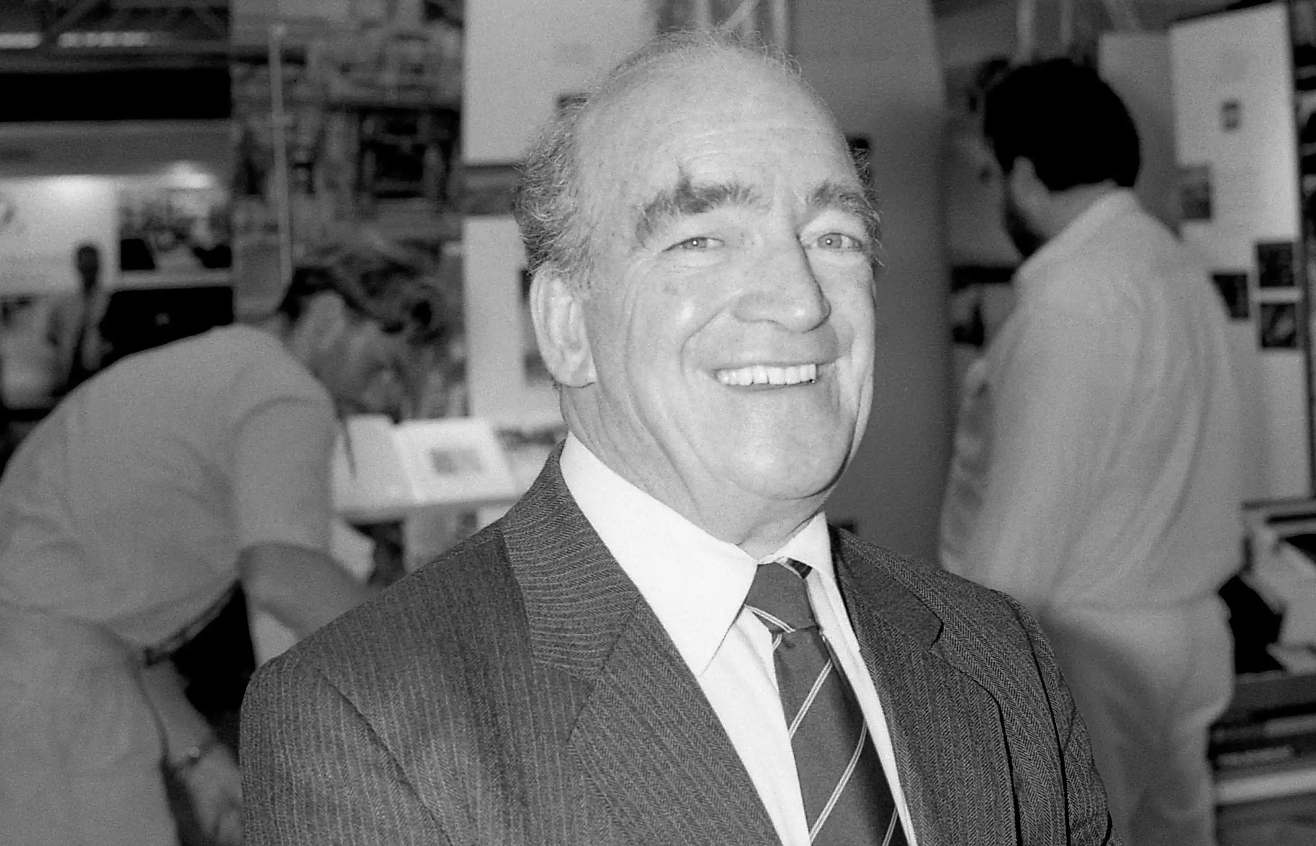 Smiling man in suit and tie, with factory workers behind him.