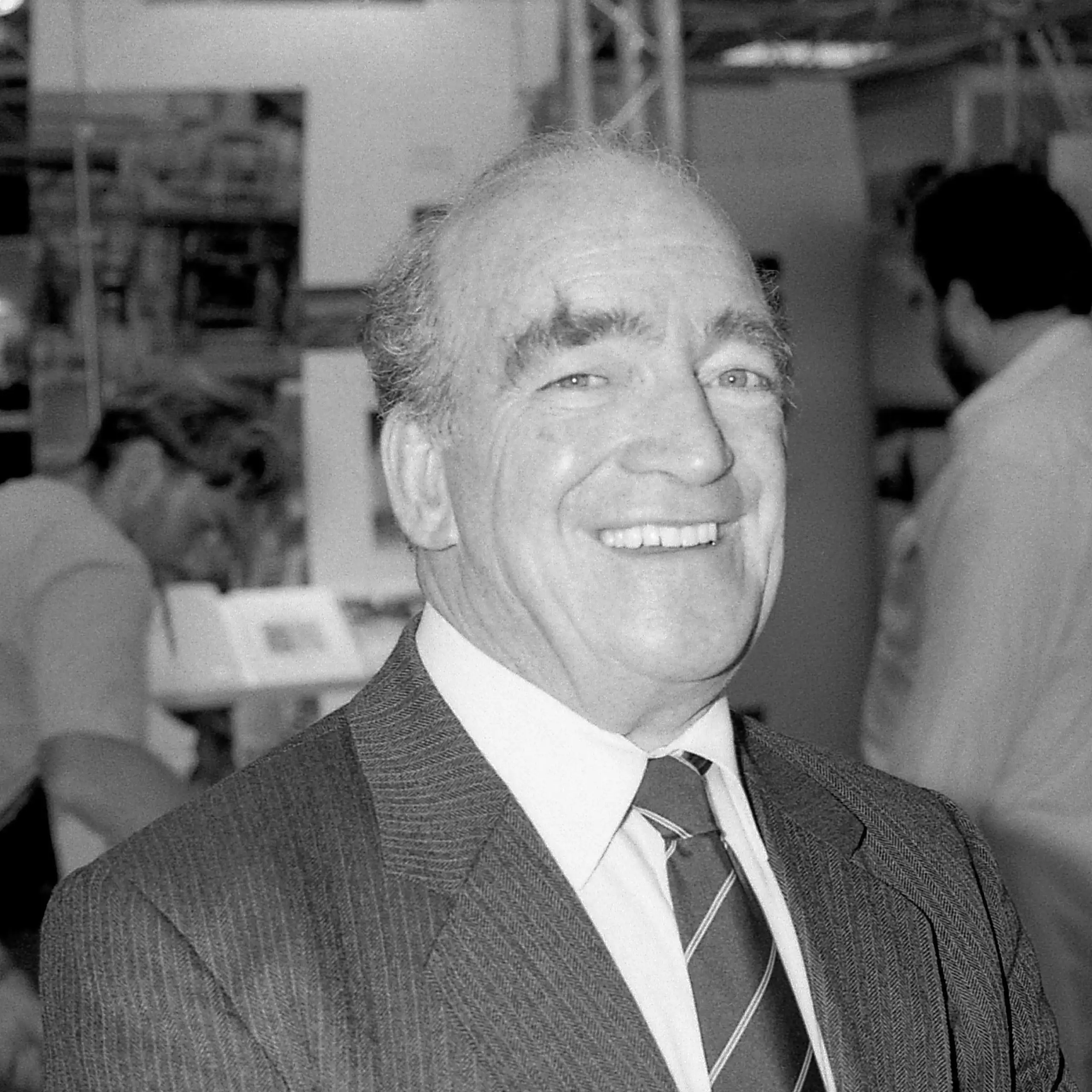 Smiling man in suit and tie, with factory workers behind him.