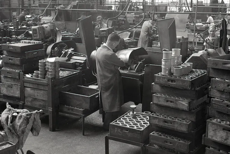 A man in a long work coat stands at a tool-making machine in a large 1950s factory
