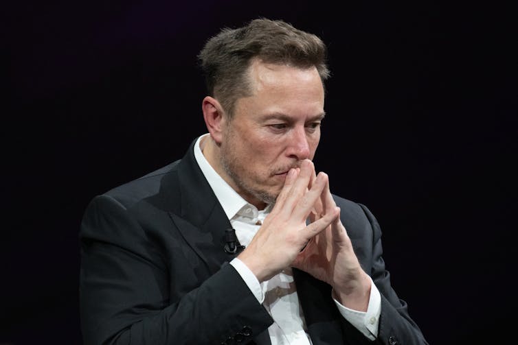 Elon Musk in a collared shirt and suit jacket, pressing his fingertips together and holding them up to his mouth