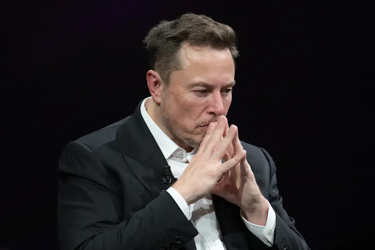 Elon Musk in a collared shirt and suit jacket, pressing his fingertips together and holding them up to his mouth