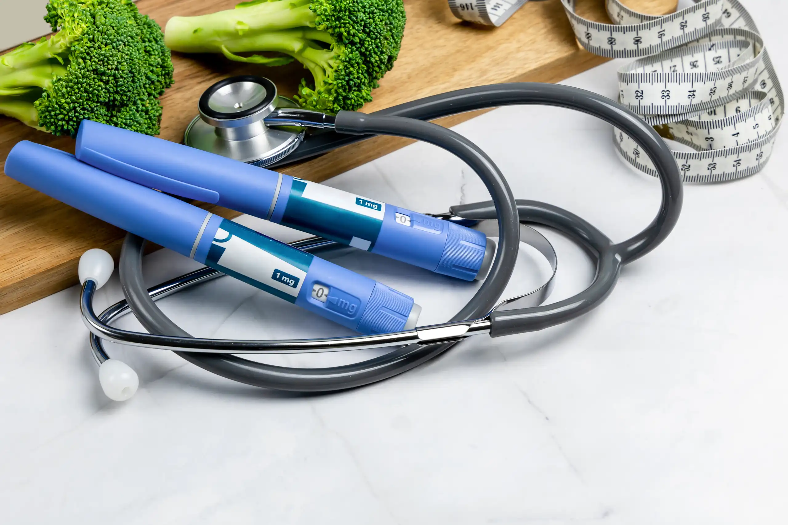 Two blue medication pens, a stethoscope, a measuring tape and some random broccoli