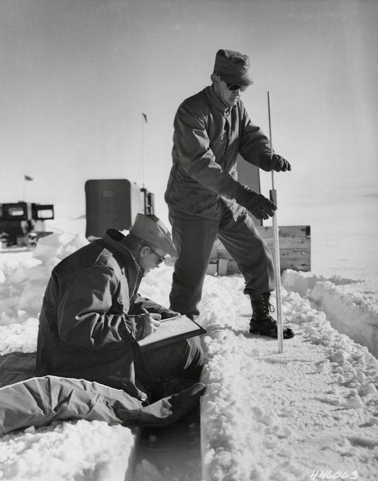 A service member pushes a thin pole into the ice to measure the thickness while another takes notes. The background is a cover of snow and ice.