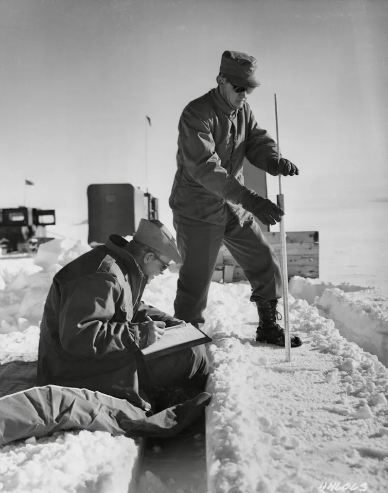 A service member pushes a thin pole into the ice to measure the thickness while another takes notes. The background is a cover of snow and ice.
