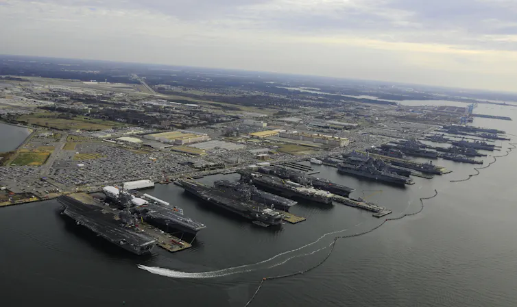 An aerial photo of more than a dozen military ships lined up at docks. The surrounding land is very flat and near sea level for miles.