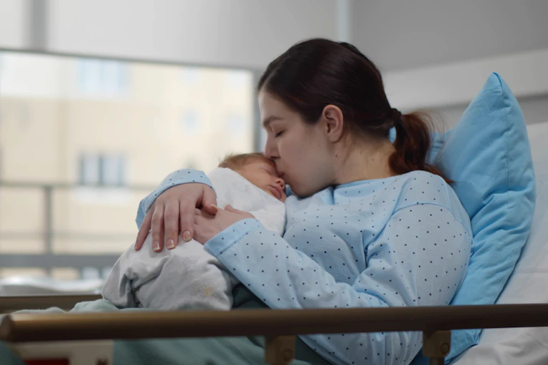 A woman in a hospital bed with a newborn, kissing its forehead