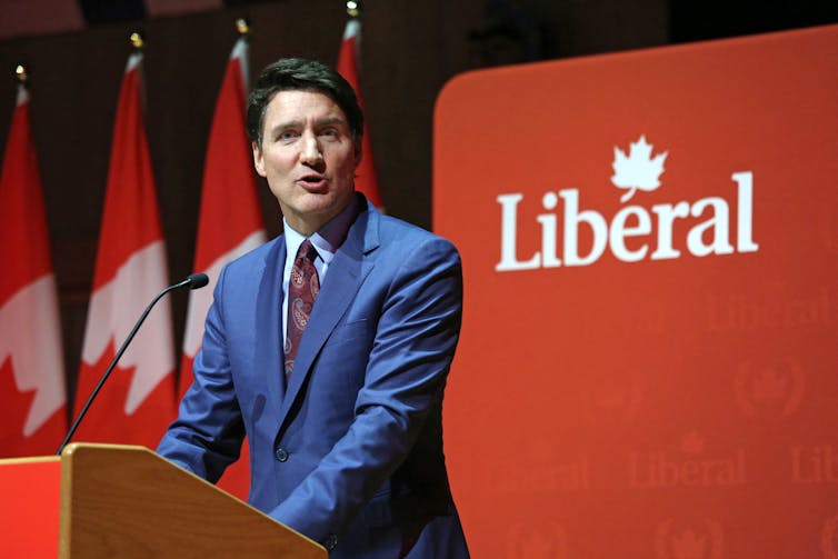 A man in a blue suit stands in front of a sign reading 'Liberal'