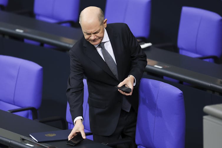 A man in a suit stands in front of blue chairs.