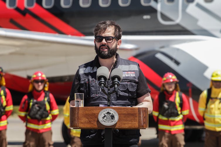 A man in dark glasses and a jacket speaks into a microphone.