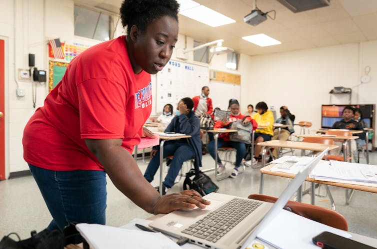 a Black woman stands while typing at a computer in a classroom with other students at desks in background