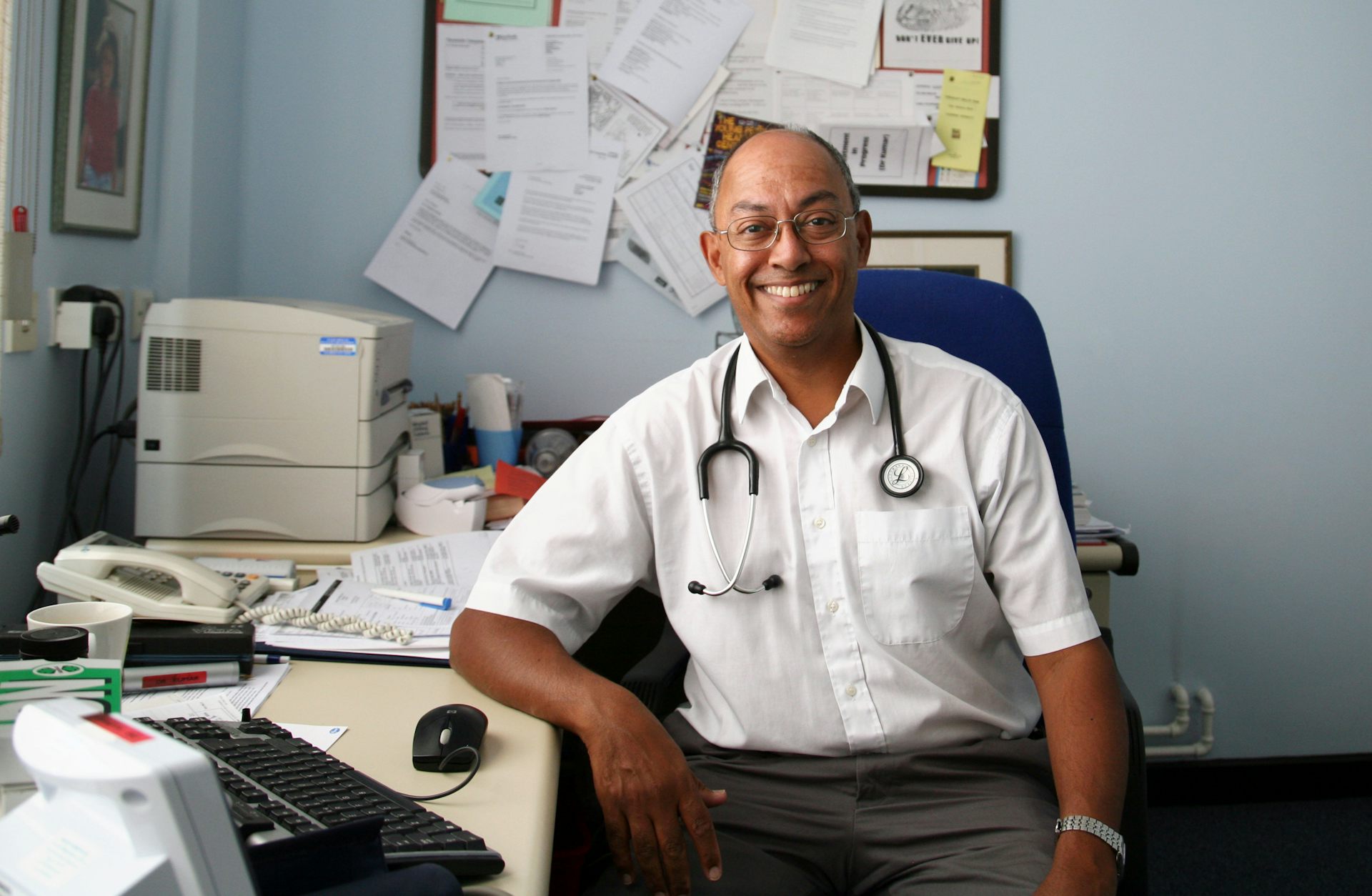 A GP in his practice, surrounded by papers, computer, printer, phone, etc.