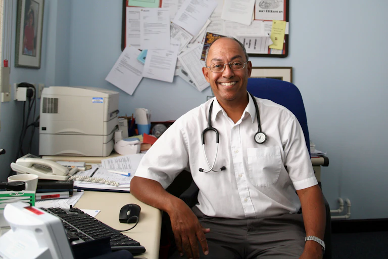 A GP in his practice, surrounded by papers, computer, printer, phone, etc.