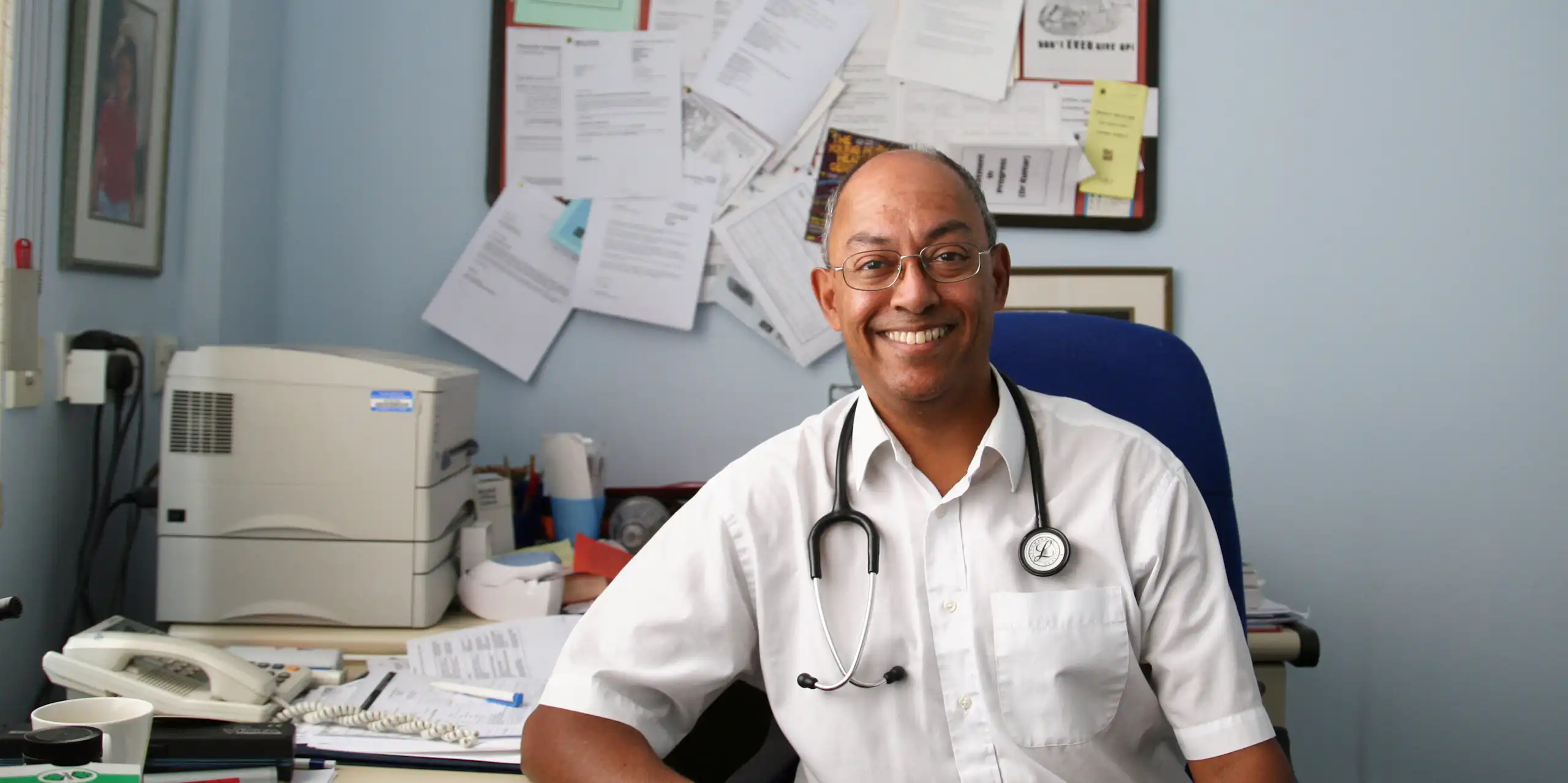 A GP in his practice, surrounded by papers, computer, printer, phone, etc.