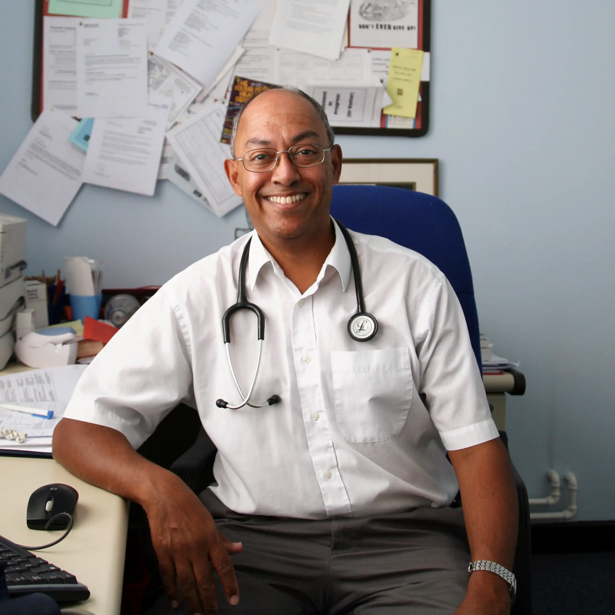 A GP in his practice, surrounded by papers, computer, printer, phone, etc.