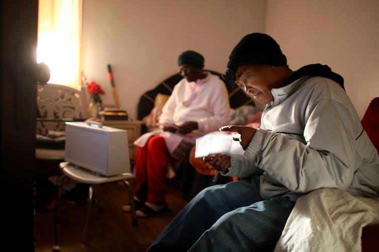 A Black woman wearing a cap and heavy coat reads a letter. In the background, an elderly woman sews