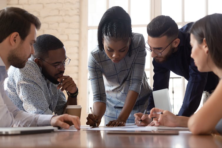 A group of people of diverse racial backgrounds sit around a conference table, looking at a document on it