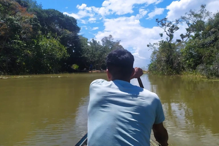 Foto mostra uma canoa sendo remada por um homem de costas. Ele está no centro do rio, e as margens dos dois lados são cobertas de florestas.