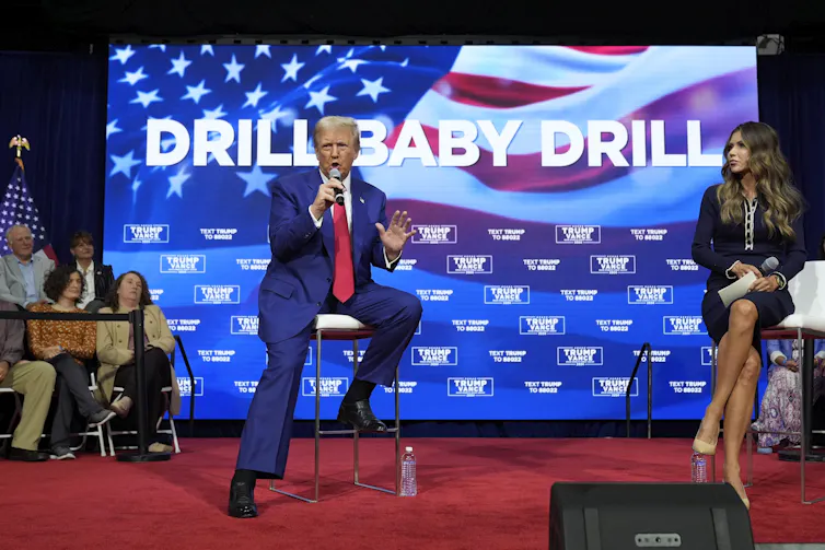 Trump sits on a stool next to South Dakota Gov. Kristi Noem on stage with a bright image behind him with the words 'drill baby, drill' and an American flag.