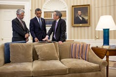 Three men stand over a couch discussing briefing material inside the White House Oval Office.
