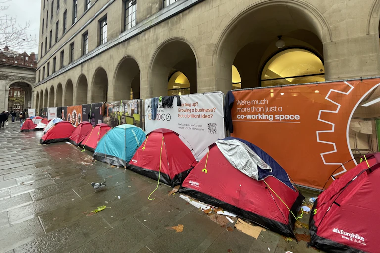 A row of mostly red camping tents in a line against the arches of a municipal building in Manchester, on a rainy day