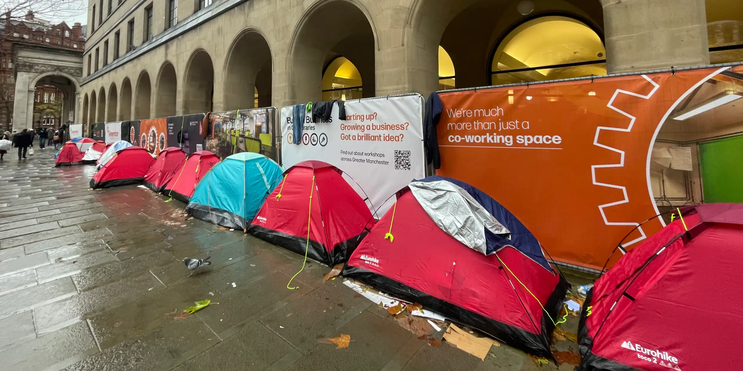 A row of mostly red camping tents in a line against the arches of a municipal building in Manchester, on a rainy day