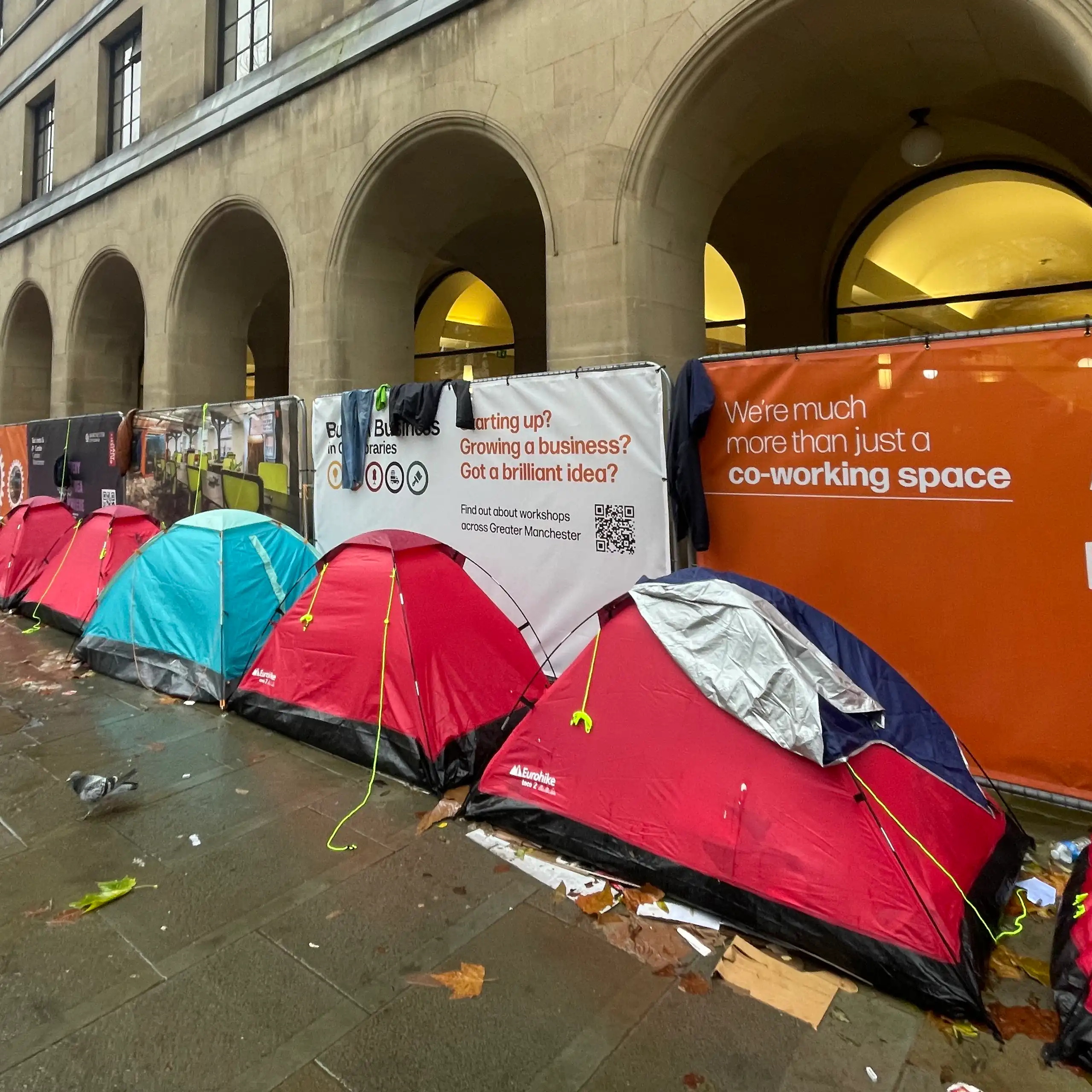 A row of mostly red camping tents in a line against the arches of a municipal building in Manchester, on a rainy day