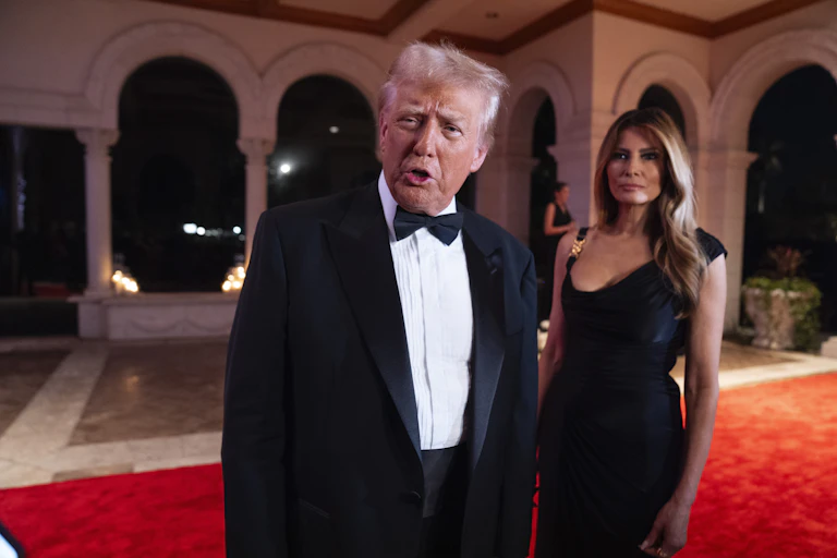 An aging man with fluffy blond-grey hair wearing a tuxedo speaks as a dark-haired woman stands behind him