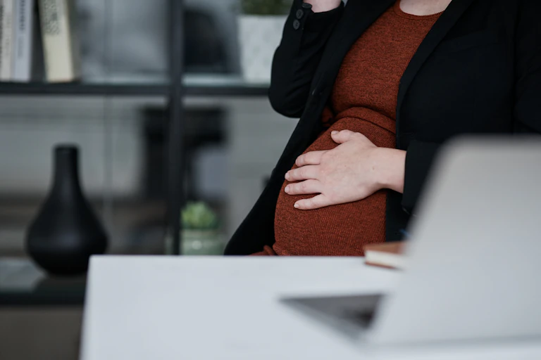 Close up of the pregnant stomach of a person sitting in front of a desk with an open laptop on it