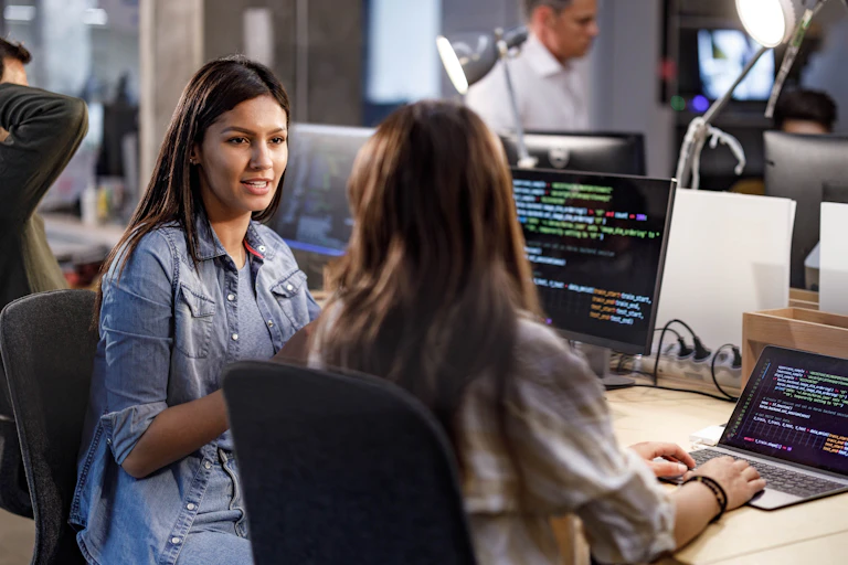 two women sit at computers and talk